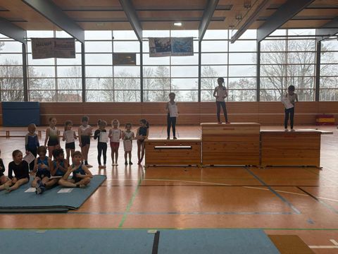Siegerehrung bei einem Kinder-Turnwettkampf in einer Sporthalle. Drei Kinder stehen auf einem Siegerpodest aus Turnkästen und halten Urkunden. Links daneben steht und sitzt eine Gruppe von etwa 15 weiteren Kindern in Turnkleidung auf dem Hallenboden und einer Turnmatte. Im Hintergrund sind große Fenster mit blattlosen Bäumen sowie Werbebanner an der Decke zu sehen.
