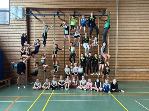 Großes Gruppenfoto von etwa 40 Kindern und Jugendlichen in einer Turnhalle. Die Gruppe hat sich pyramidenförmig an und auf einer Sprossenwand mit Kletterstangen arrangiert – einige hängen oben an den Stangen, andere klettern seitlich, weitere stehen in der Mitte und die jüngsten Kinder sitzen unten auf dem grünen Hallenboden. Viele tragen schwarz-grüne Turnanzüge, andere bunte Sportkleidung. Alle blicken fröhlich in die Kamera.