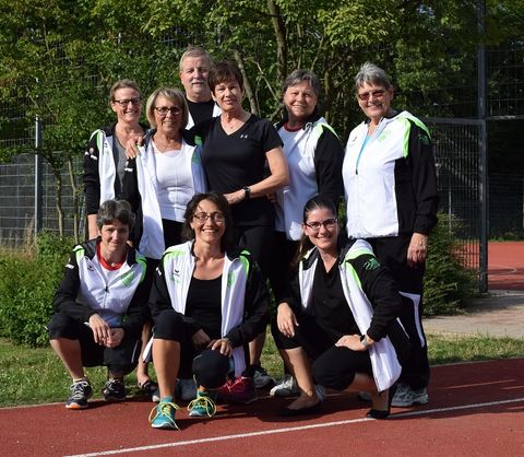 Gruppenfoto von zehn Personen auf einer Leichtathletik-Tartanbahn im Freien, im Hintergrund grüne Bäume und ein Zaun. Die Gruppe besteht aus neun Frauen und einem Mann, die meisten tragen schwarz-weiße Trainingsjacken mit grünen Akzenten. Sechs Personen stehen in der hinteren Reihe, drei knien vorne. Alle lächeln in die Kamera.