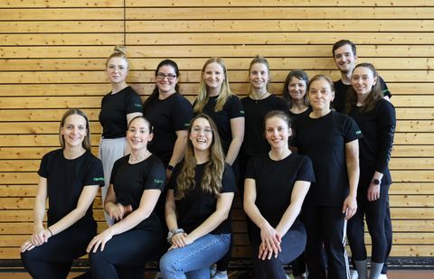 Gruppenfoto von zwölf Personen in einer Turnhalle vor einer Holzsprossenwand. Alle tragen schwarze T-Shirts mit grünem „Trainer"-Schriftzug. Die Gruppe besteht aus elf Frauen und einem Mann, aufgestellt in zwei Reihen – vier Personen sitzen vorne, acht stehen dahinter. Alle lächeln in die Kamera.