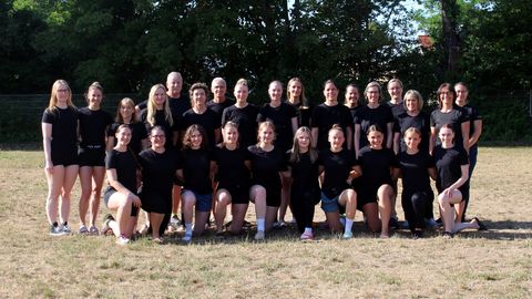 Großes Gruppenfoto von etwa 30 Personen in einer Turnhalle vor einer Holzsprossenwand. Alle tragen schwarze T-Shirts, teilweise mit grünem „Trainer"-Schriftzug. Die Gruppe ist in drei Reihen aufgestellt – die vordere Reihe kniet oder hockt, die mittlere und hintere Reihe steht. Die Gruppe besteht überwiegend aus Frauen verschiedenen Alters sowie zwei Männern. Alle lächeln in die Kamera.