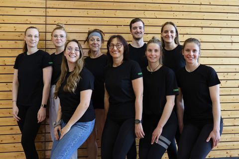 Gruppenfoto von neun Personen in einer Turnhalle vor einer Holzsprossenwand. Alle tragen schwarze T-Shirts, einige mit grünem „Trainer"-Schriftzug. Die Gruppe besteht aus acht Frauen und einem Mann verschiedenen Alters, locker in zwei Reihen aufgestellt. Alle lächeln in die Kamera.